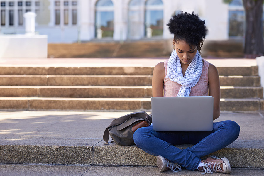 young college student sitting outside legs crossed on a step working on a laptop
