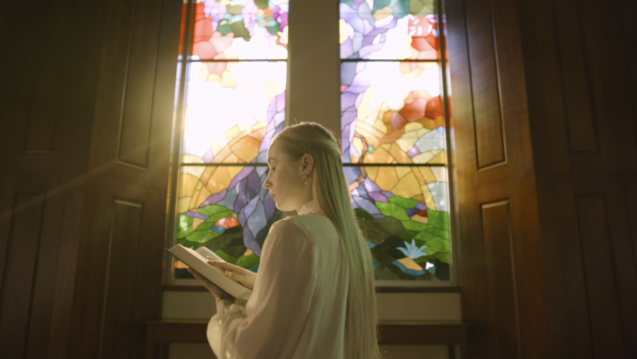 Young woman holding a Bible, reading it, standing in front of backlit stained glass.
