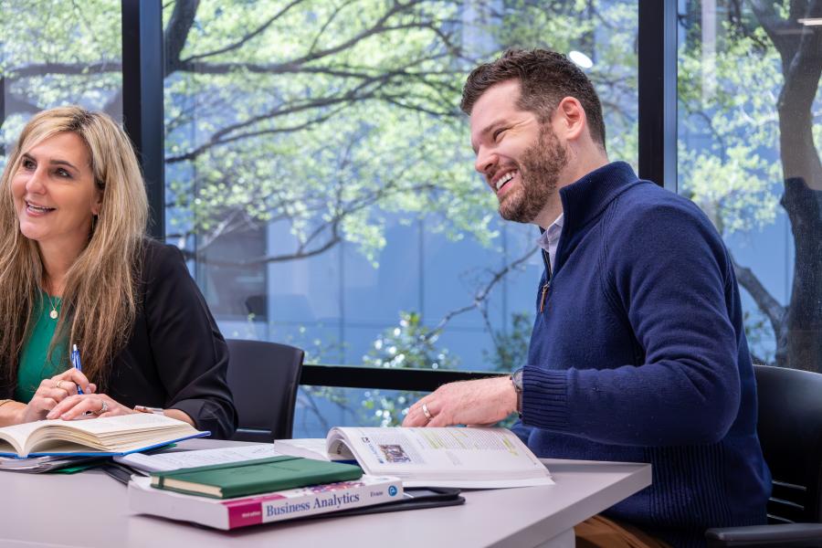 MBA students around a table with textbooks smiling and trees in the window outside behind them